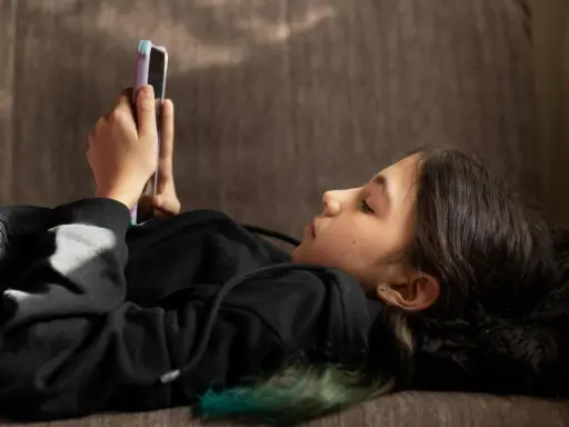 Ein Mädchen liegt auf einer Couch und schaut auf ihr Smartphone. Sie ist in ein schwarzes Kapuzensweatshirt gekleidet. Dies spiegelt aktuelle Trends der Jugend wider.