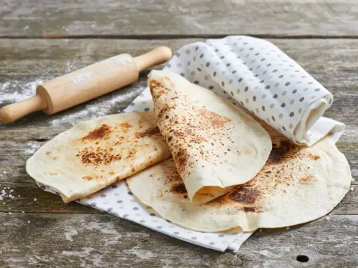 Świeżo usmażona tortilla z patelni, lekko przypieczona, leży na lnianej ściereczce obok wałka.