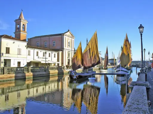 Barche a vela tradizionali ormeggiate sul canale di Cesenatico mare, con edifici storici e riflessi sull'acqua.