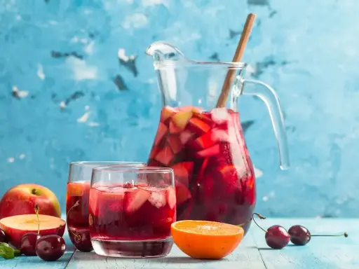 En karaff med röd bål, fylld med frukt och is, står bredvid två glas. Äpplen, körsbär och en apelsinskiva dekorerar bordet. Ett perfekt bål recept för sommaren.