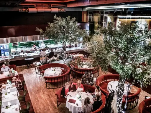 Elegant restaurang med runda, röda bås och träd. Köket syns i bakgrunden. Många gäster njuter av sin måltid. L'avventura recensioner verkar positiva.