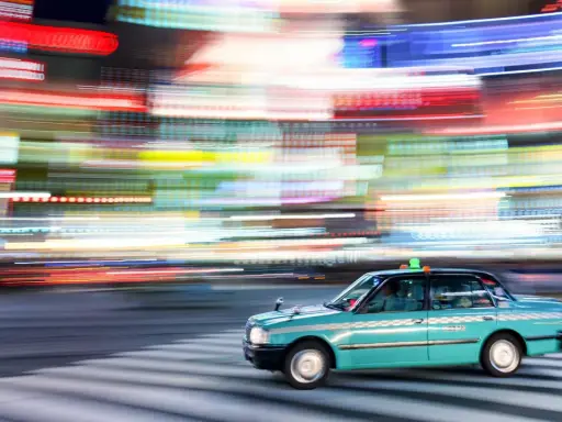 Un taxi azzurro sfreccia su strisce pedonali, con luci sfocate di città che creano un effetto dinamico, perfetto per imparare come fare foto in movimento.