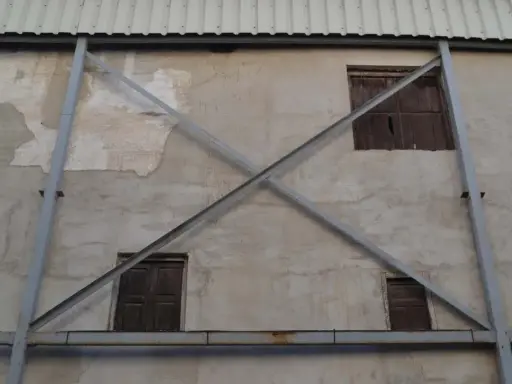 Estructura metálica en forma de X que refuerza una pared de concreto con ventanas de madera. La riostra, en este caso, es un elemento de soporte crucial.