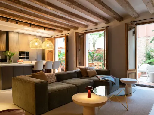 Salón moderno con vigas de madera vistas, sofá modular, cocina integrada y acceso a patio. Ideal para poner falso techo y unificar espacios.