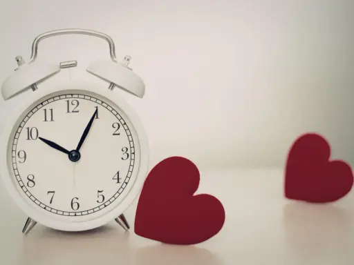 Biały budzik pokazuje 10:08. Obok niego leżą dwa czerwone serca. 0505 znaczenie - czas na miłość.