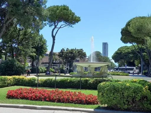Fontana z wodą w **Parco Fellini Rimini**, otoczona zielenią i kwiatami. W tle widać autobus i wieżowiec.