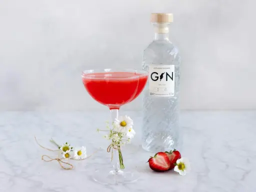 En röd drink i ett coupeglas, bredvid en flaska gin från Gotlands Ginfabrik. Jordgubbar och små blommor dekorerar bordet.