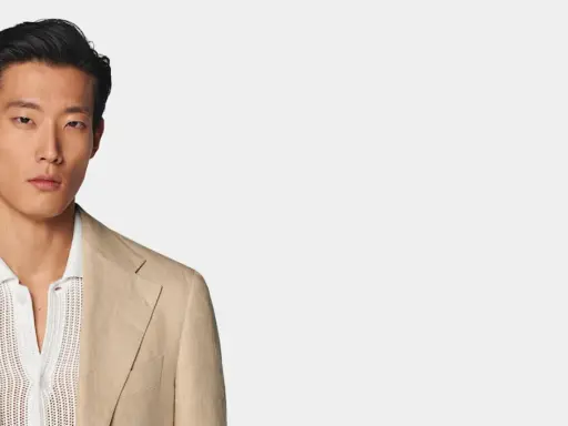 A man models a tan linen blazer over a white textured polo shirt, perfect for a smart casual dress code for a wedding.