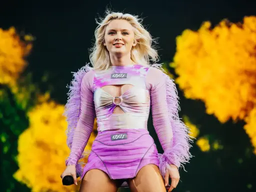 Zara Larsson konsert. Hon står på scenen i en lila och rosa outfit med fjädrar på ärmarna.