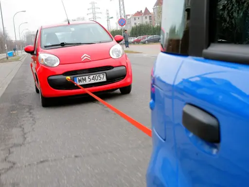 Czerwony Citroen holowany przez niebieski samochód. Pomarańczowa linka holownicza jest napięta, sugerując ruch i prędkość.
