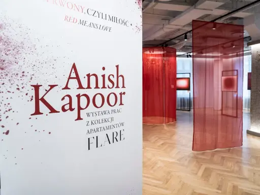 Biały plakat z napisem "Anish Kapoor" i czerwonymi rozpryskami na tle wystawy z czerwonymi zasłonami.