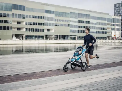 Ein Mann joggt mit Kinderwagen an einer modernen Fassade entlang. Das Kind schläft friedlich im Wagen.
