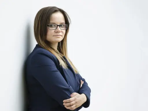 Kobieta w okularach i granatowej marynarce stoi oparta o ścianę, ze skrzyżowanymi rękami.