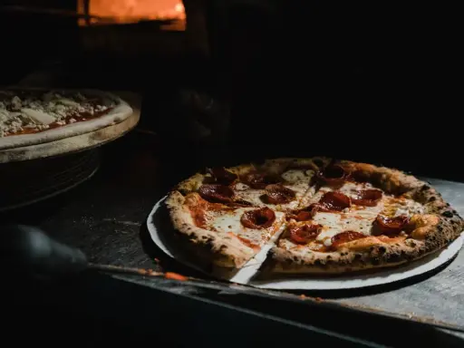 Upieczona pizza pepperoni na talerzu, obok surowa pizza w piecu.