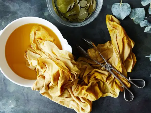 A yellow fabric is soaking in a bowl of golden liquid, part of a natural dye process. Eucalyptus leaves are nearby.