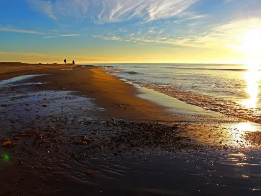 Två personer promenerar längs stranden vid Skagens udde i solnedgången. Vågorna sköljer in mot land.