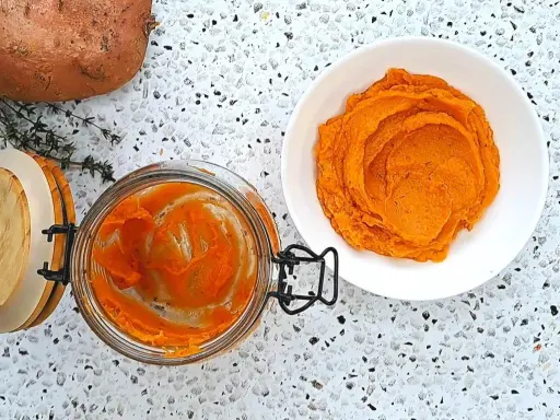 Pomarańczowe purée z batatów w słoiku i na talerzu, obok surowy batat i gałązka tymianku.