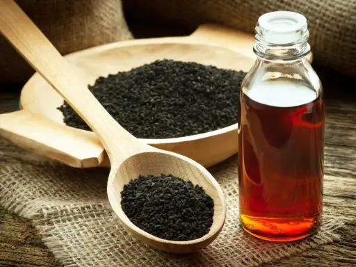 Huile de nigelle ambrée dans un flacon, à côté de graines noires dans une cuillère en bois. Découvrez les bienfaits de l'huile de nigelle.