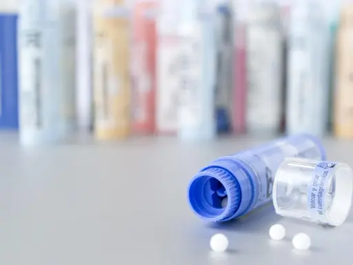 Flacons de granules homéopathiques, certains en arrière-plan floutés. Un tube bleu ouvert laisse échapper quelques billes blanches. Une solution naturelle pour la dermite séborrhéique, comme une huile essentielle, pourrait être contenue dans ces remèdes.