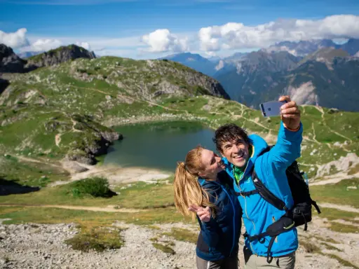 Coppia felice si scatta un selfie con il lago Coldai sullo sfondo, circondato da montagne verdi e un cielo azzurro con nuvole bianche.