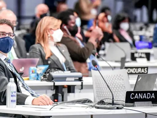 Teilnehmer der Klimakonferenz, darunter ein Mann mit Brille und Maske, sitzen an Tischen mit Laptops und Namensschildern wie "CANADA".