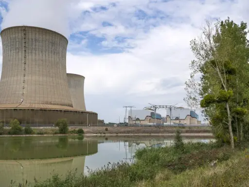 Kühltürme eines Atomkraftwerks in Frankreich, die Dampf abgeben. Ein ruhiger See spiegelt die Anlage wider, während am Ufer Bäume und Gras wachsen.