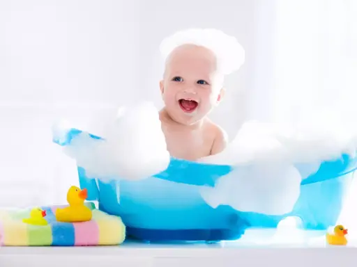 Uśmiechnięty noworodek w pianie, podczas kąpieli. Obok kolorowy ręcznik i gumowe kaczuszki. Pielęgnacja noworodka to radość.