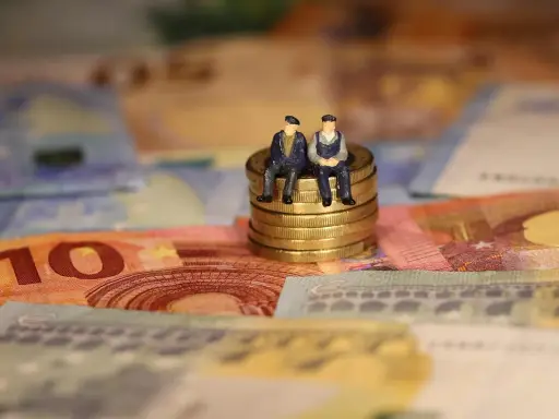 Dos figuritas sentadas en monedas de oro sobre billetes de euros. Reflexionan: como puedo cotizar los años que me faltan.
