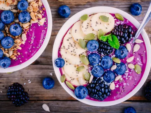 Zwei Schalen mit gesunden Bowls, gefüllt mit lila Smoothie, garniert mit Blaubeeren, Bananenscheiben, Brombeeren, Kürbiskernen und Granola.