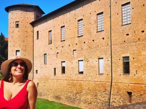 Donna sorridente con cappello di paglia davanti a un castello di mattoni rossi. Un'idea per cosa vedere a Piacenza in un giorno.