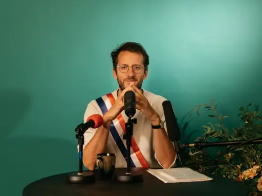 Un homme avec une écharpe tricolore, prêt à parler. Il est entouré de micros, peut-être pour un discours plein de langue de bois.