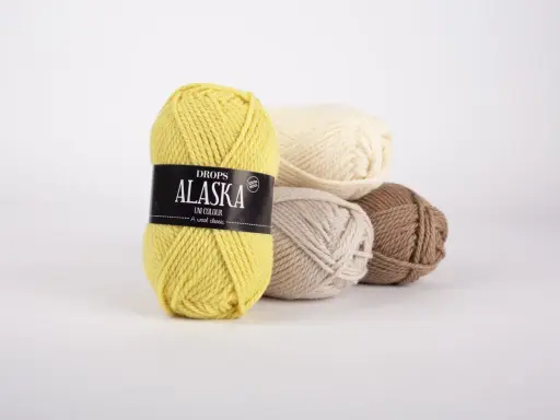 Gula, beige, ljusbruna och vita nystan av Drops Alaska garn ligger i en hög.