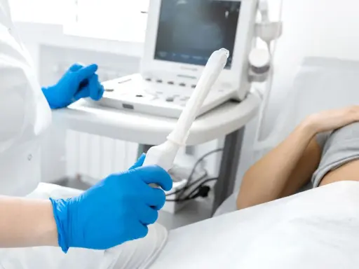 Medico con guanti blu tiene sonda per ecografia transvaginale. Paziente sdraiata sullo sfondo.