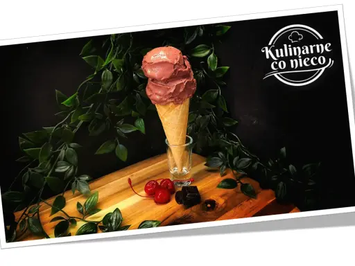 Dwa kule czekoladowych lodów białkowych w wafelku, ozdobione wiśniami i kawałkami gorzkiej czekolady.