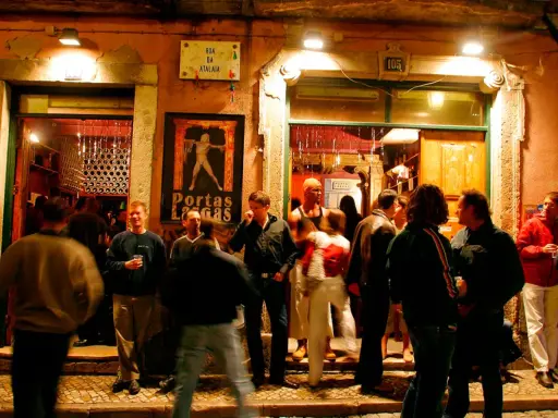Menschen versammeln sich vor einem beleuchteten Lokal in Bairro Alto. Ein Mann hält ein Glas, andere unterhalten sich.