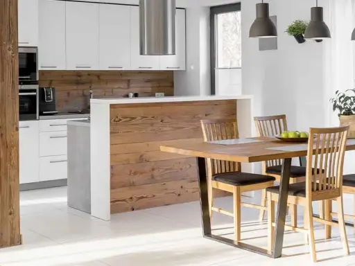 Cocina semiabierta al salón con barra de madera, encimeras blancas y electrodomésticos integrados. Mesa de comedor de madera con sillas.