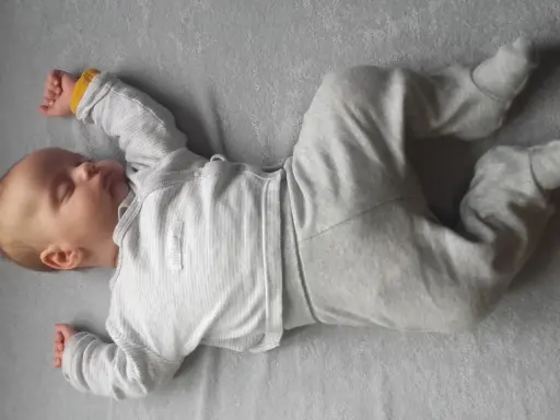 Śpiący noworodek w szarym pajacyku. Jego rączki i nóżki są ułożone w lekkiej asymetrii, co jest naturalne dla niemowląt.