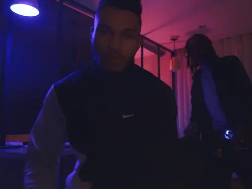 Dwóch mężczyzn w ciemnym pomieszczeniu, oświetlonym niebieskim i różowym światłem. Jeden z nich, 730 huncho, ma na sobie bluzę z logo Nike.