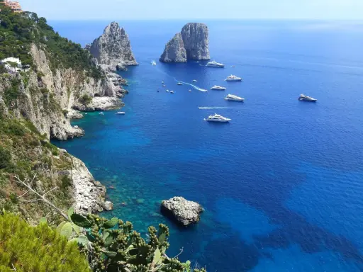 Scogliere verdi e rocce imponenti si affacciano sul mare blu intenso, con yacht che navigano. Un'immagine che ispira su come raggiungere Capri da Napoli.