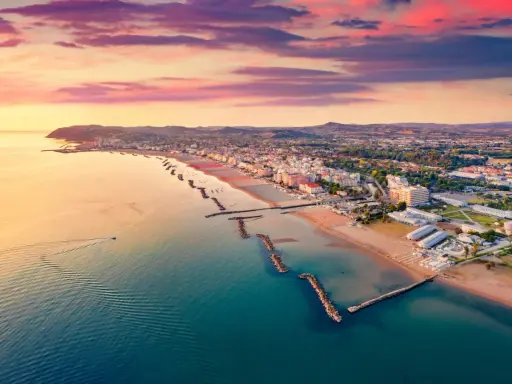Alba rosa sul cattolica mare, con la costa e le colline sullo sfondo. Una barca solca le acque calme.