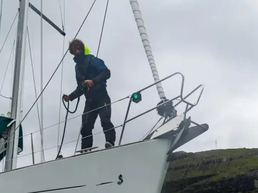 Un marinier, les amarres avis, manœuvre sur le pont d'un voilier blanc, face à une côte rocheuse verdoyante.