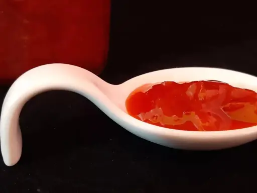 Une cuillère blanche présente une portion de confiture fraise rhubarbe, d'un rouge vif et appétissant. Un pot de confiture est visible en arrière-plan.