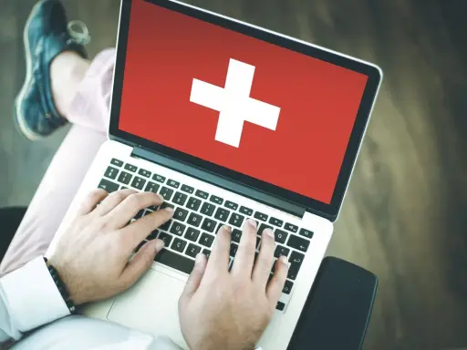 Osoba pisząca na laptopie z flagą Szwajcarii na ekranie.