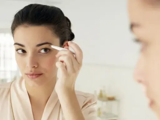 Une jeune femme ajuste ses sourcils broussailleux avec une pince à épiler devant un miroir, dans une tenue de chambre satinée.