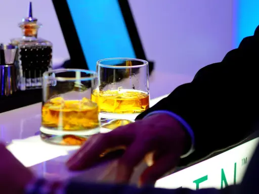 Dwa kieliszki złocistej whisky na barze, idealne na dzień whisky. Dłoń w garniturze sięga po drinka.