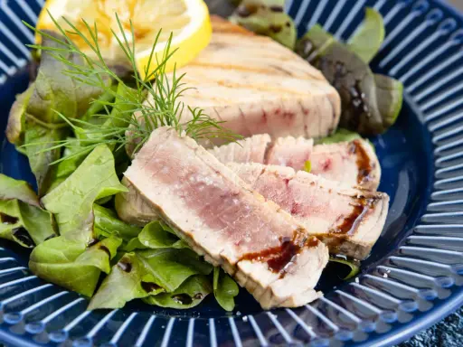 Ein perfekt gegrilltes Thunfischsteak, in Scheiben geschnitten, serviert mit frischem Salat, Zitrone und Dill auf einem blauen Teller.
