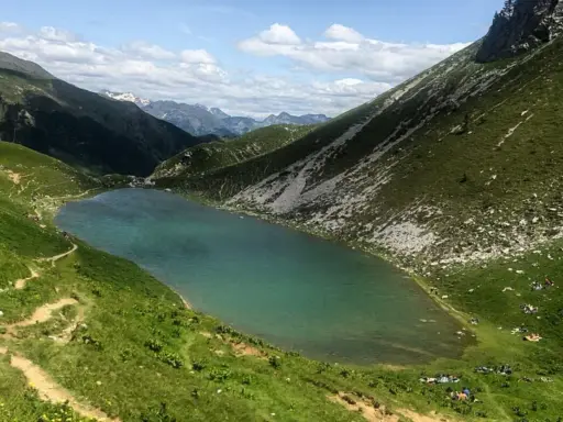 Un sentiero serpeggia tra prati verdi verso il lago Branchino, le cui acque turchesi riflettono le montagne circostanti. Alcune persone si godono un picnic sulle rive.