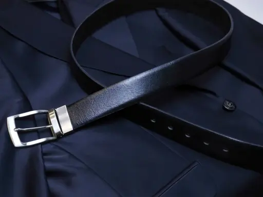 A black leather belt with a silver buckle rests on a dark navy suit jacket. Do you wear a belt with a tuxedo? This image shows a classic accessory.