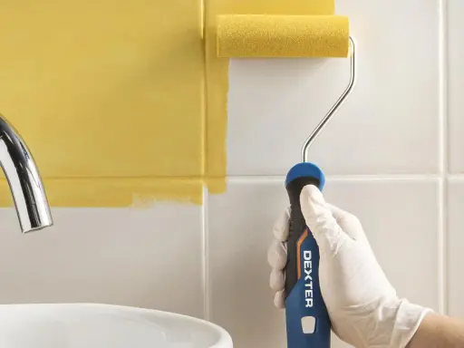 Mano con guante blanco pintando azulejos de cocina de blanco a amarillo con rodillo.