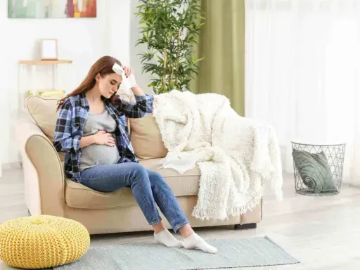 Donna incinta seduta sul divano con un fazzoletto sulla fronte, che soffre di raffreddore in gravidanza.